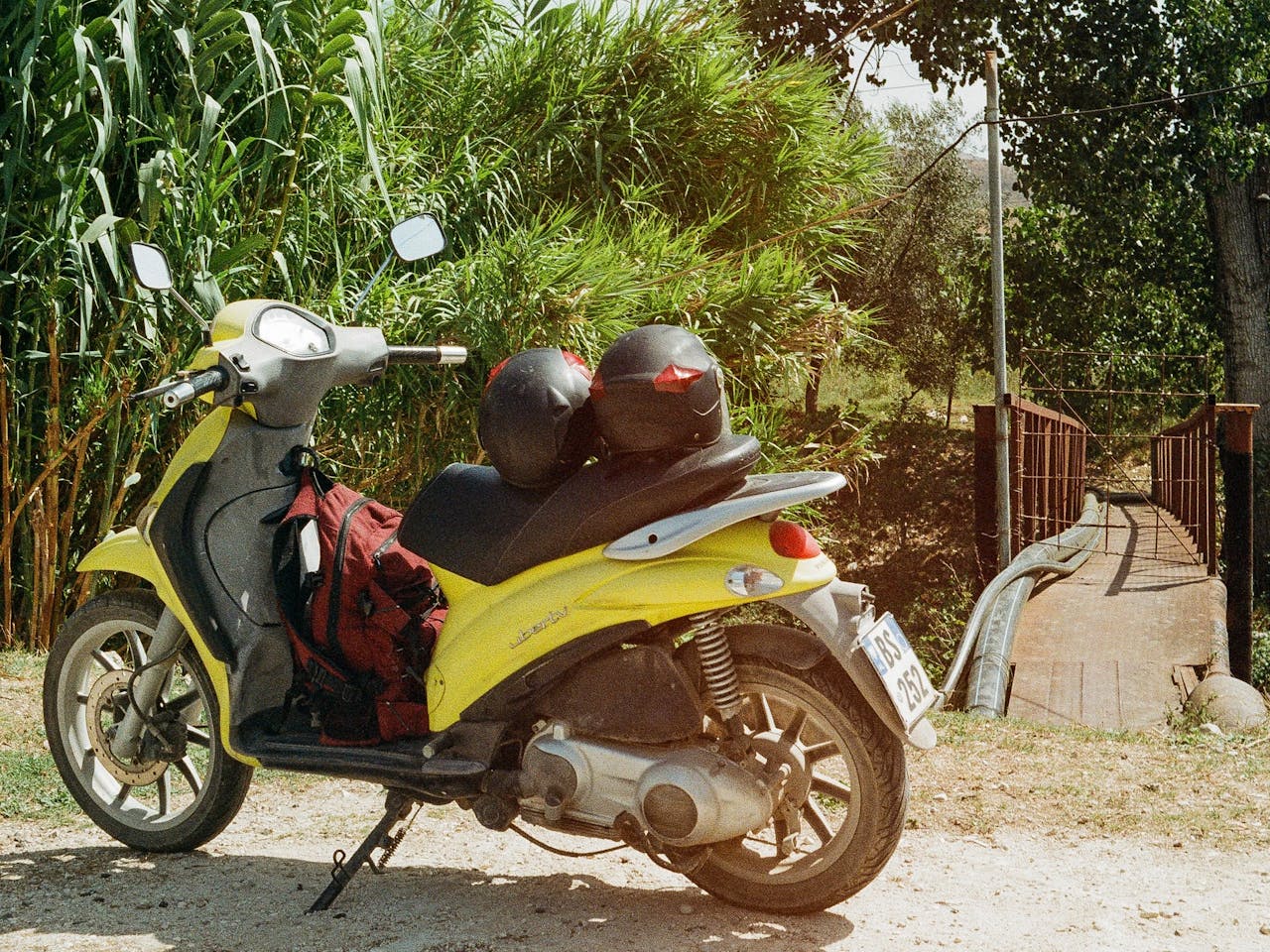 一辆亮黄色踏板车，配有头盔和包，停放在户外环境中的一座质朴桥旁。